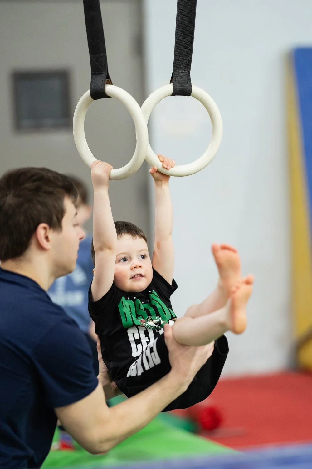 A young boy hanging from gymnastic rings while an adult supports him, in a gym setting.
