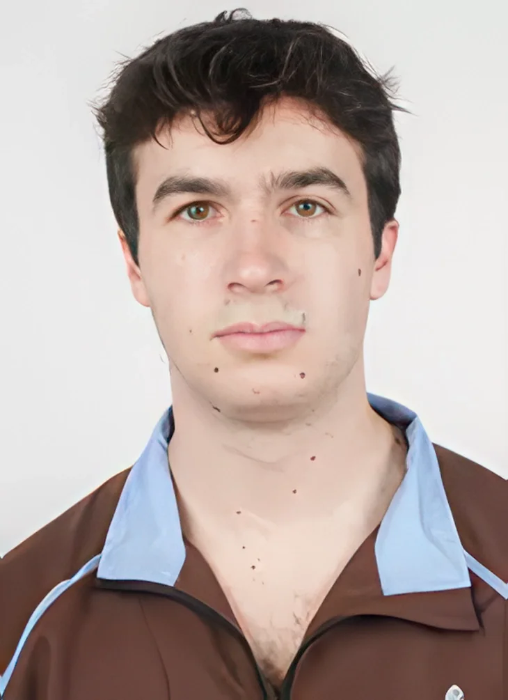 A young man with dark hair and light skin, wearing a brown and blue sports jacket, looking directly at the camera with a neutral expression.