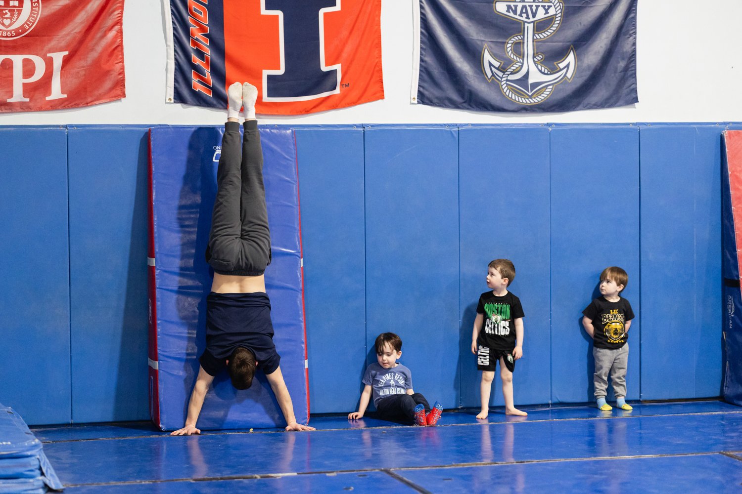 A man performing a handstand against a padded wall in a gym with three children watching. The gym has blue padded walls and flags hanging overhead.