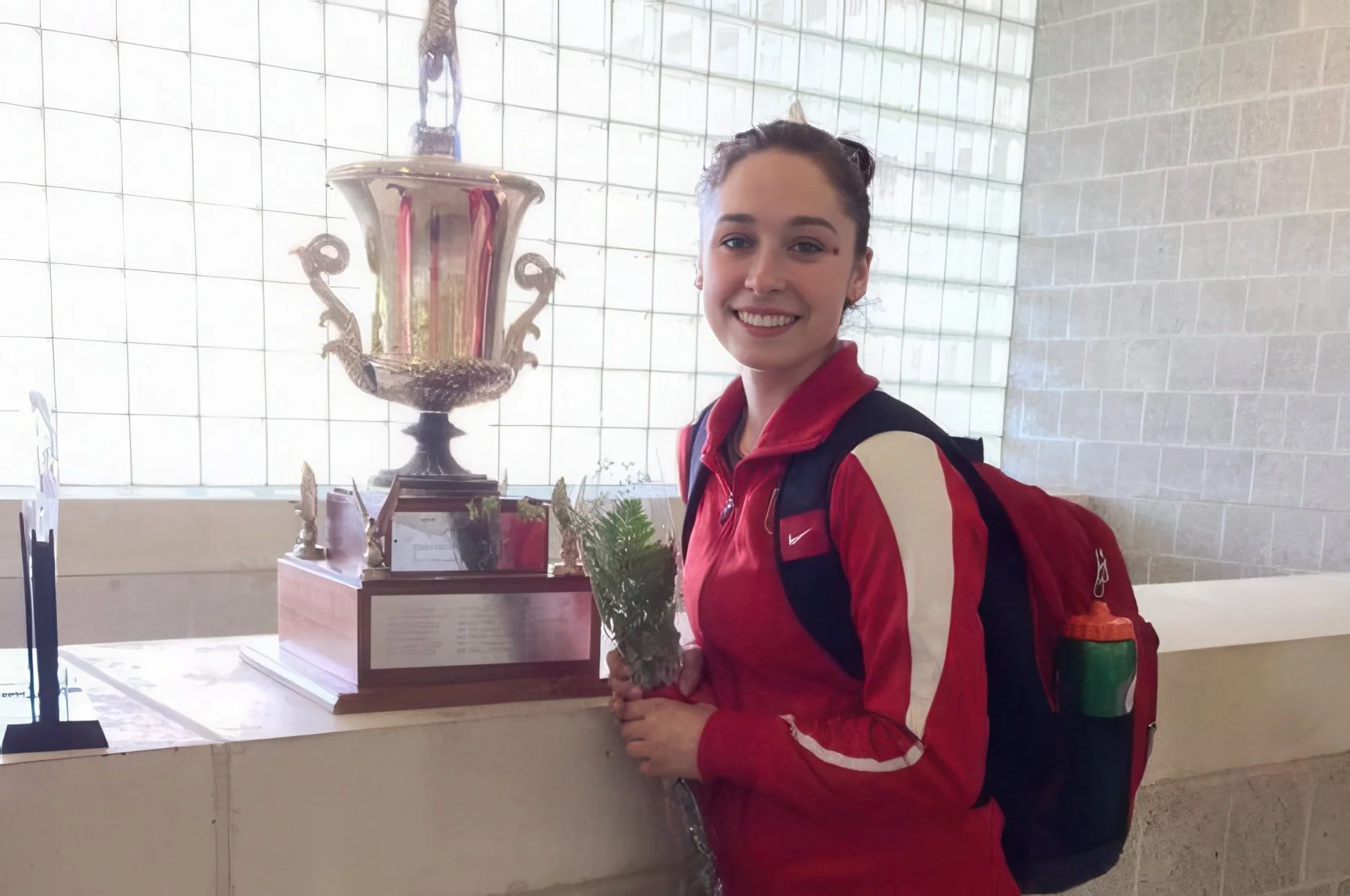 A young woman with a red and beige jacket and a backpack smiling and holding a small arrangement of greenery, standing next to a large ornate trophy on display.