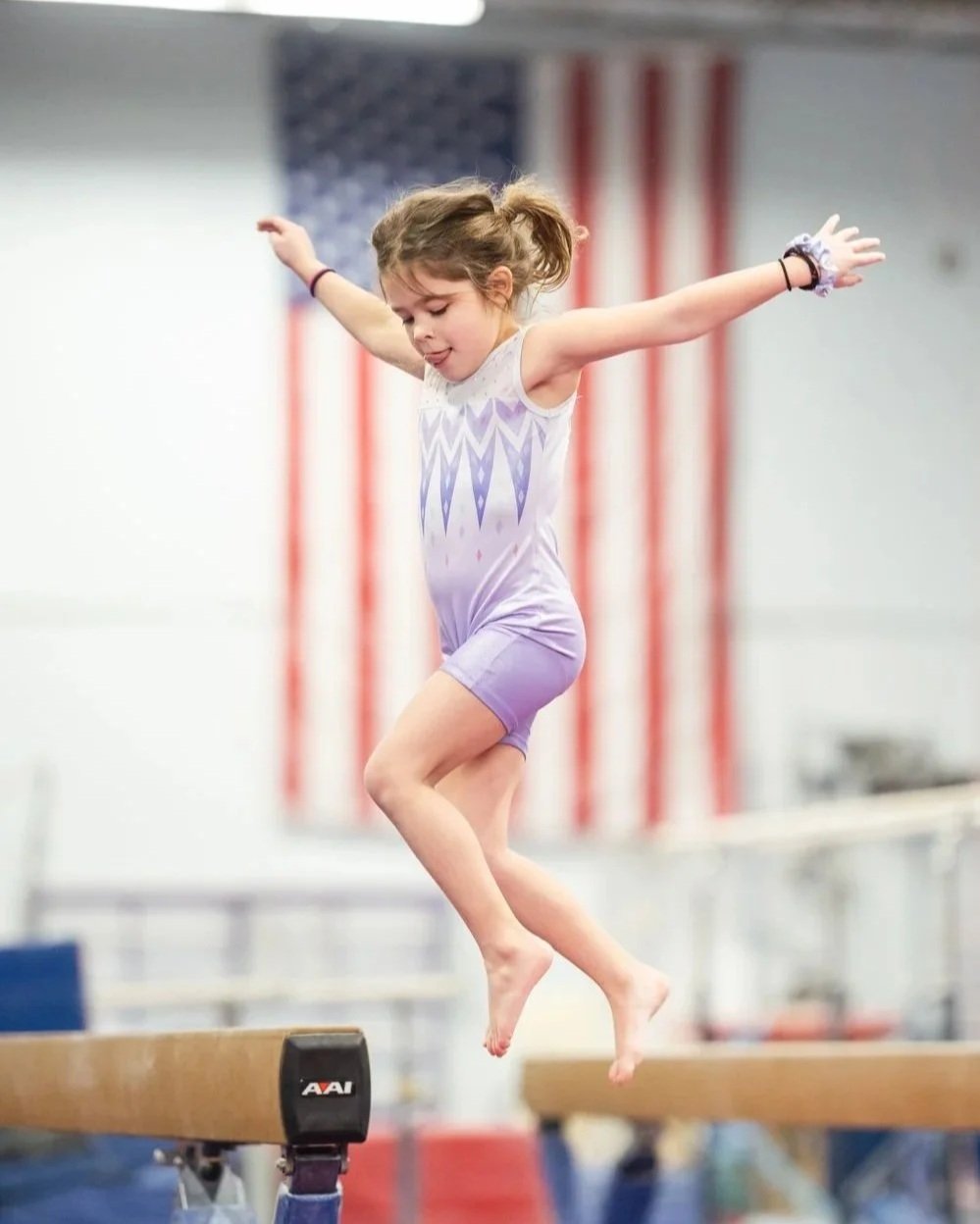 Young girl in a purple gymnastics leotard jumping on a balance beam in a gymnasium with American flags in the background.