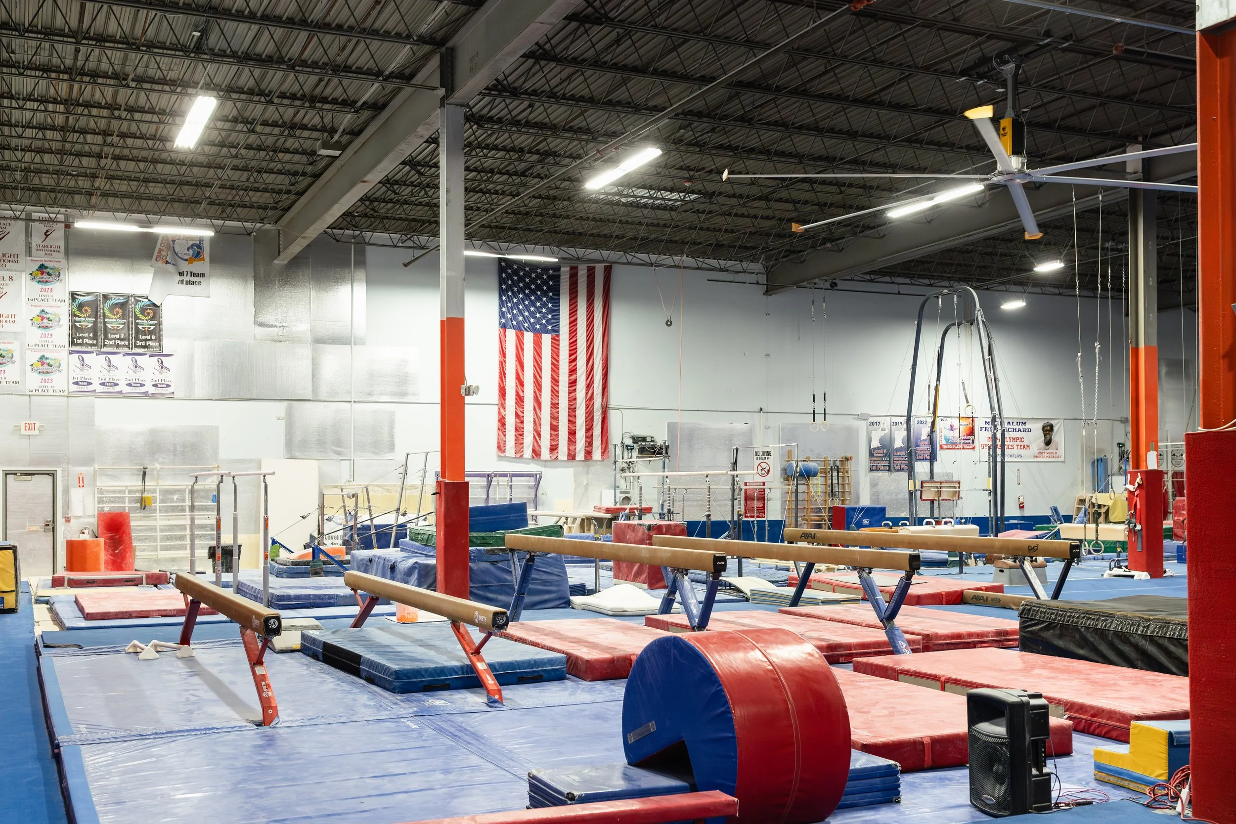 Interior of a gymnastics training facility with mats, balance beams, and gymnastics equipment, American flag hanging on the wall, banners and posters, high ceiling with industrial lighting.