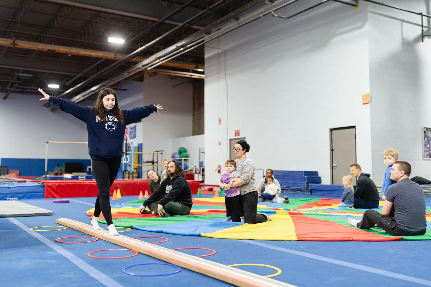 A young girl with long brown hair balancing on a wooden beam with her arms outstretched inside a gymnasium. She is practicing gymnastics or balance exercises. Several adults and children are seated on a colorful mat in the background, observing or wa