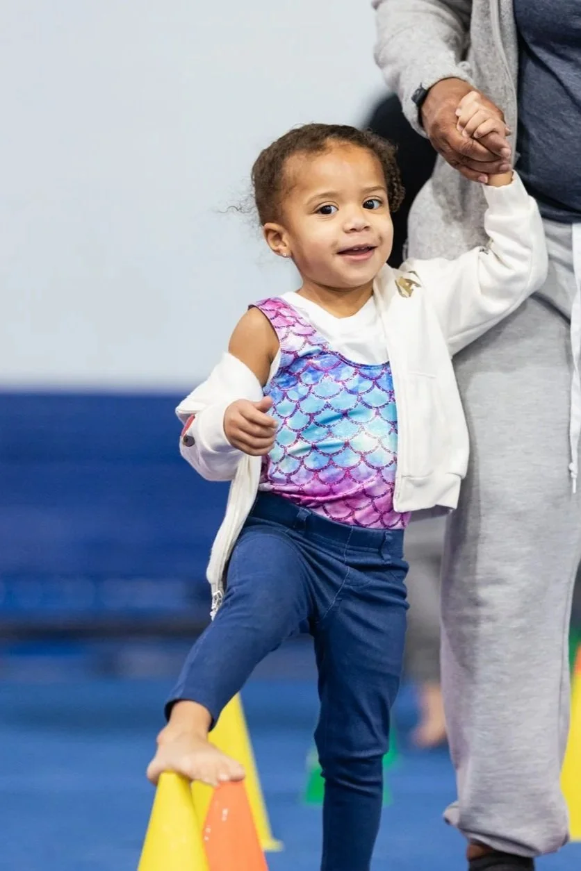 Young girl in a colorful top and navy pants balancing on a yellow cone, holding an adult's hand for support during an activity.