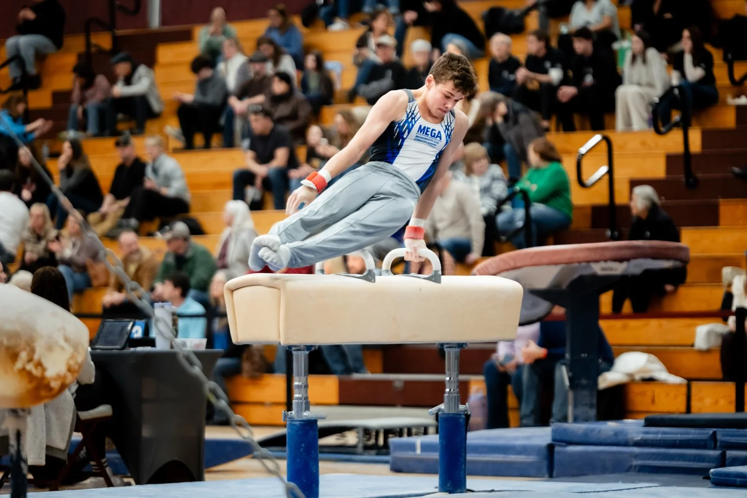 Young male gymnast in a blue and white uniform performing a routine on the rings in an indoor gymnasium with an audience seated on wooden bleachers in the background.