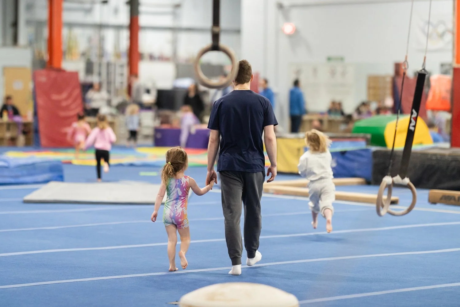 A man walks hand-in-hand with two young girls inside an indoor gymnastics gym. The background shows gymnastic equipment, mats, and other children playing.
