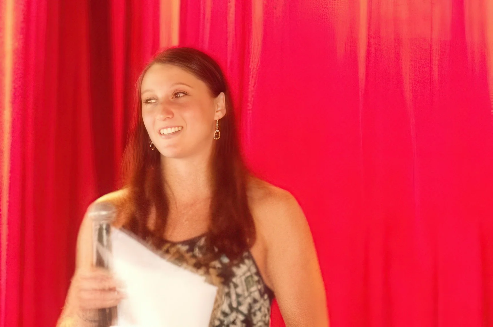 A young woman with brown hair wearing earrings, smiling and holding a drink, standing in front of a red curtain.