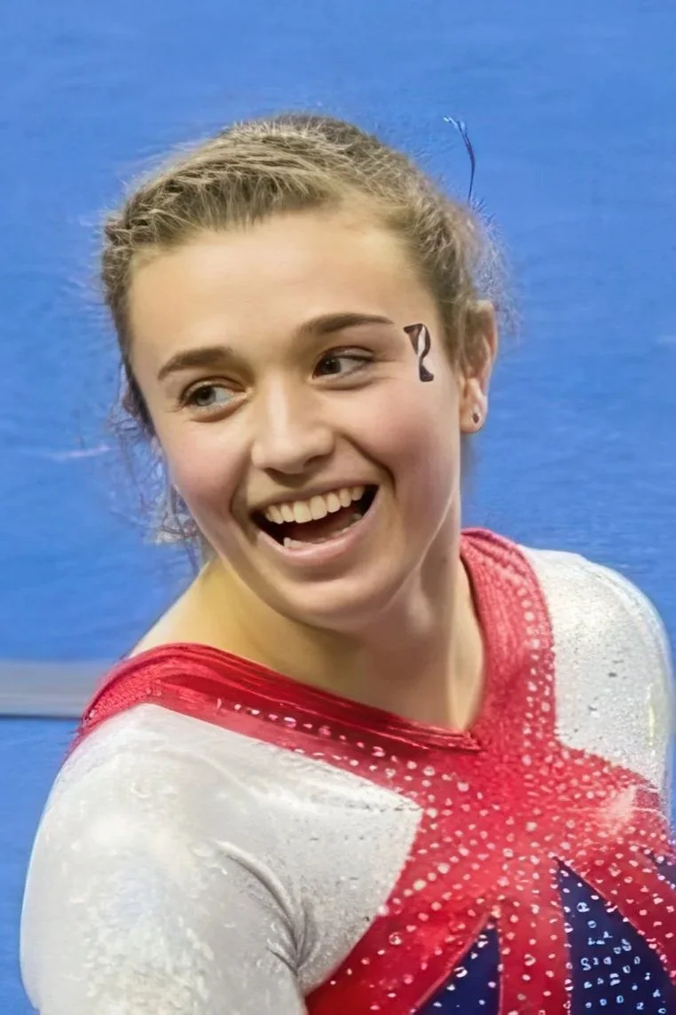 A young female gymnast with a painted question mark on her cheek, smiling in front of a blue wall, wearing a red, white, and blue leotard.