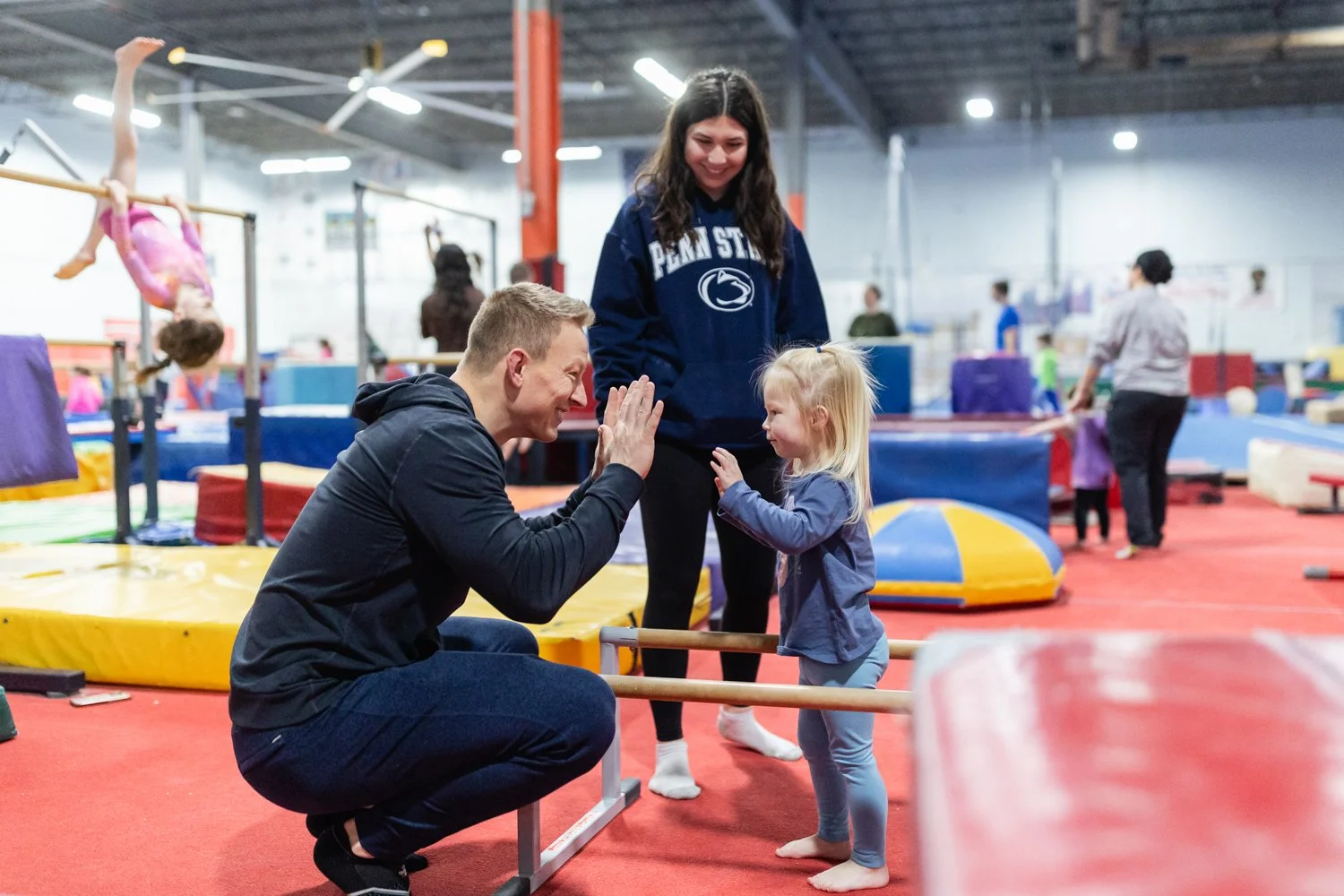 A man and a little girl are giving each other high fives at an indoor gymnastics facility, with a woman and other children visible in the background.