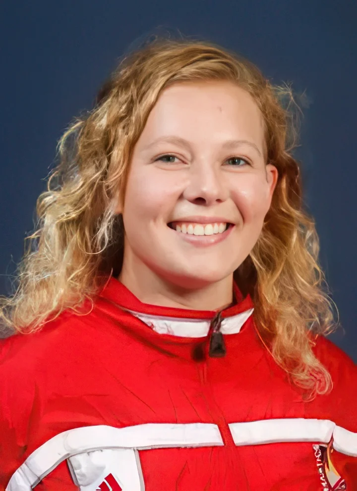 A young woman with curly blonde hair wearing a red athletic jacket with white accents, smiling at the camera against a blue background.