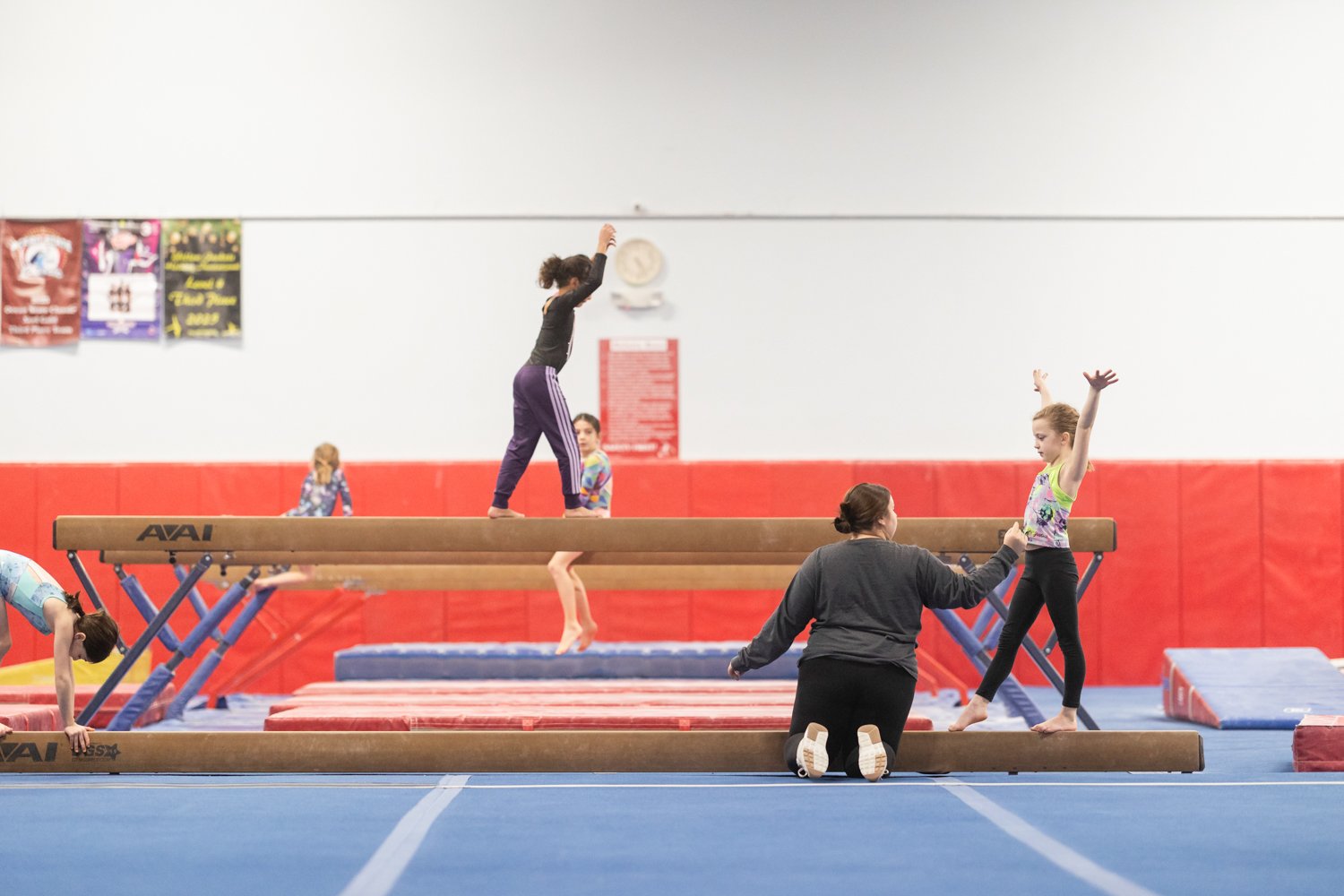 Indoor gymnastics training with children practicing balances and movements on balance beams, supervised by an instructor.