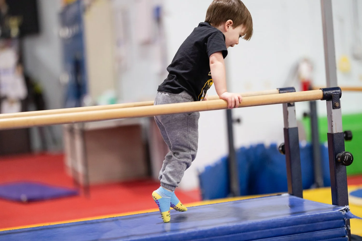 A young boy in gray sweatpants and a black t-shirt balancing on a padded gym beam with gray and yellow socks, inside a gym or physical therapy center.