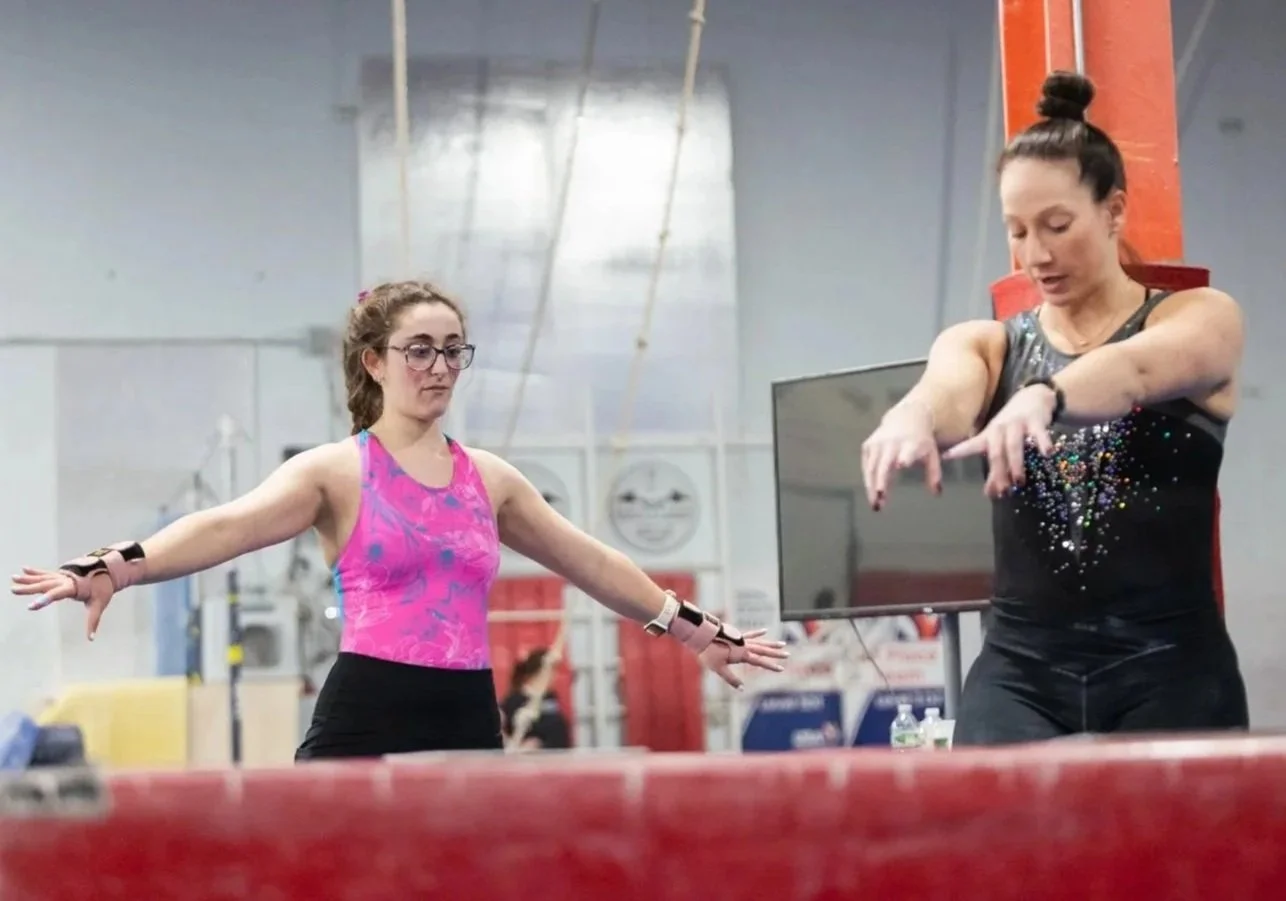Two women standing in a gym, one with arms extended to the sides, wearing a pink and purple sports top and black shorts, and the other with arms forward, wearing a black tank top with colorful embellishments and black workout pants, preparing for gymnastics or fitness training.