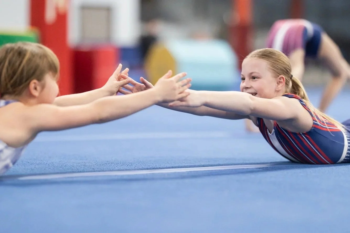 Two young girls performing a fitness exercise on a blue mat, reaching out to each other while lying face down, with other children visible in the background.