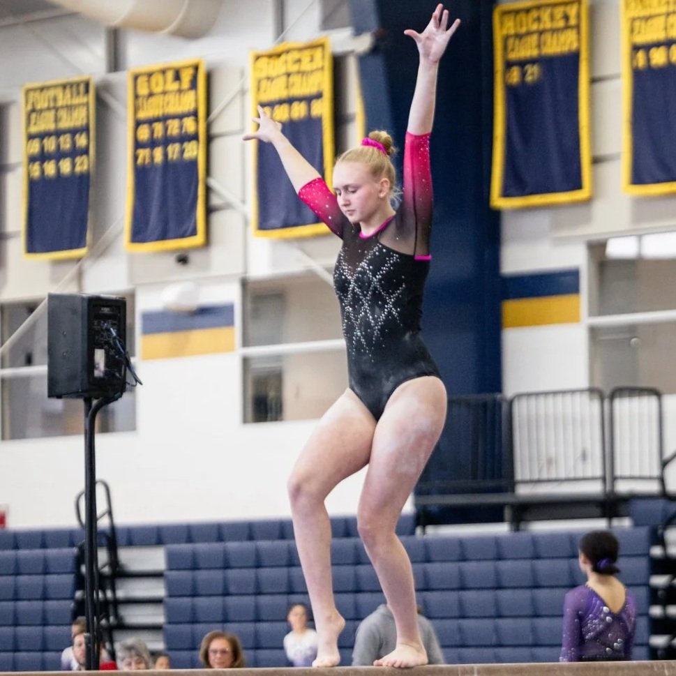 A young female gymnast performing on the balance beam at a gymnastics competition, with spectators watching in the background.