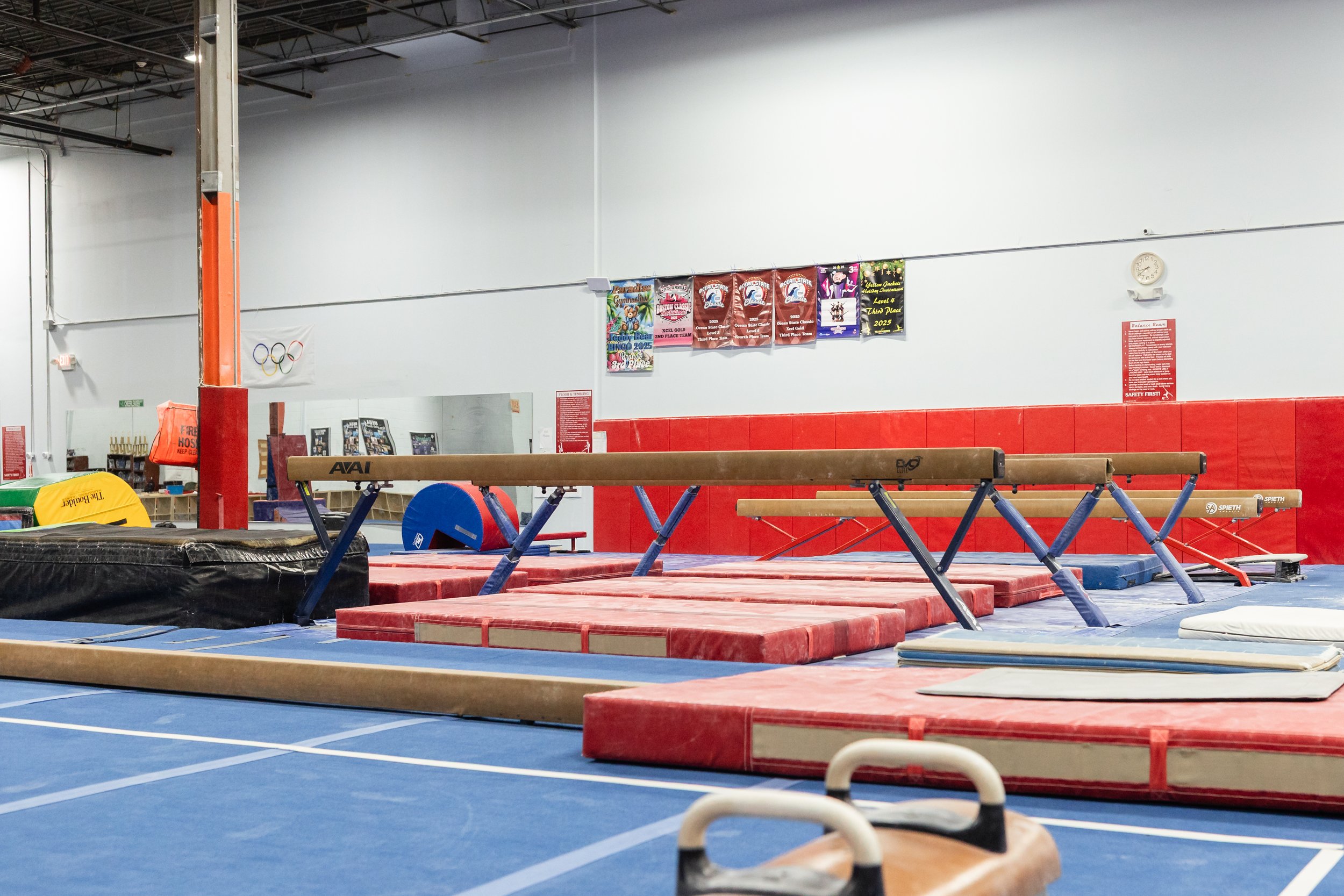 An empty gymnastics training area with balance beams, padded mats, and various gymnastics equipment in a spacious indoor gymnasium