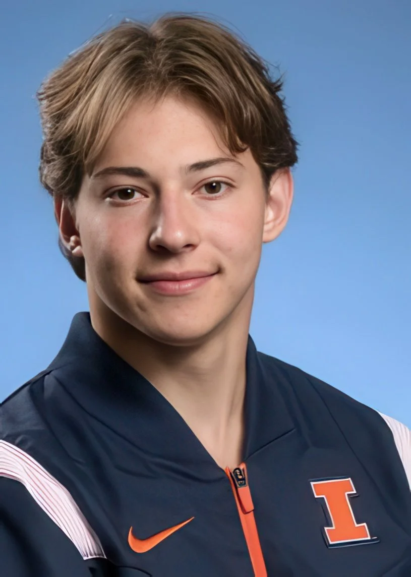 A young man with light skin and brown hair, wearing a navy blue athletic jacket with an orange Nike logo and a letter 'I' on it, smiling softly against a blue background.