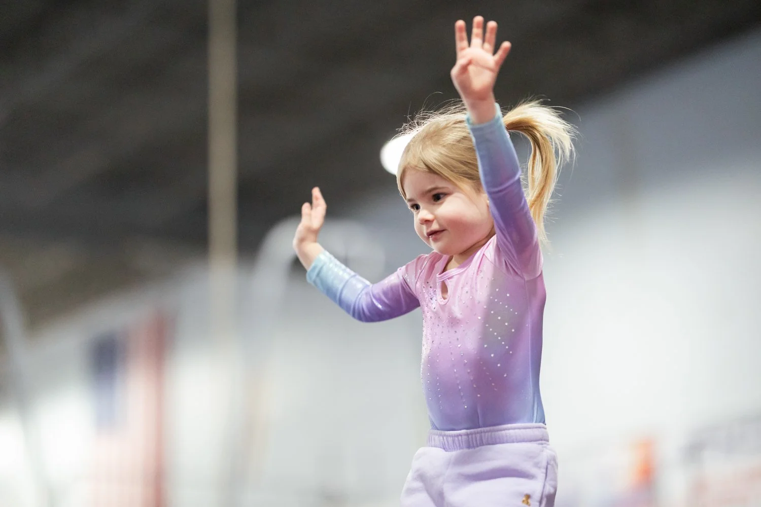 A young girl with blonde hair tied in a ponytail wearing a sparkly purple gymnastics leotard and white sweatpants, practicing or performing a move with her arms raised in an indoor gym or training facility.