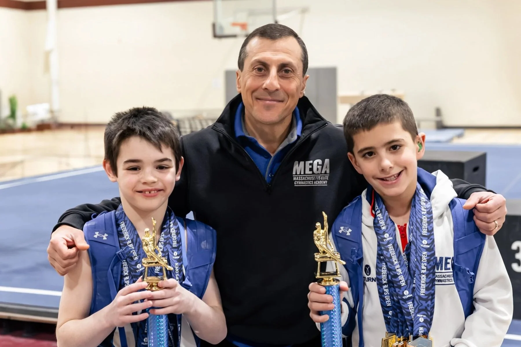 Two young boys and an adult male coach at a gymnastics competition holding trophies and medals, standing on a gymnastics mat.
