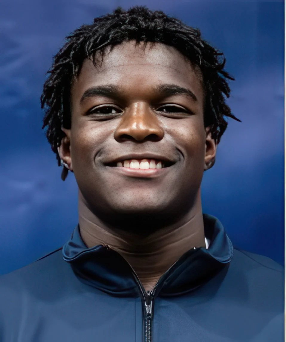 Close-up of a young man with short, curly black hair, smiling, wearing a dark athletic jacket, against a cloudy blue sky background.