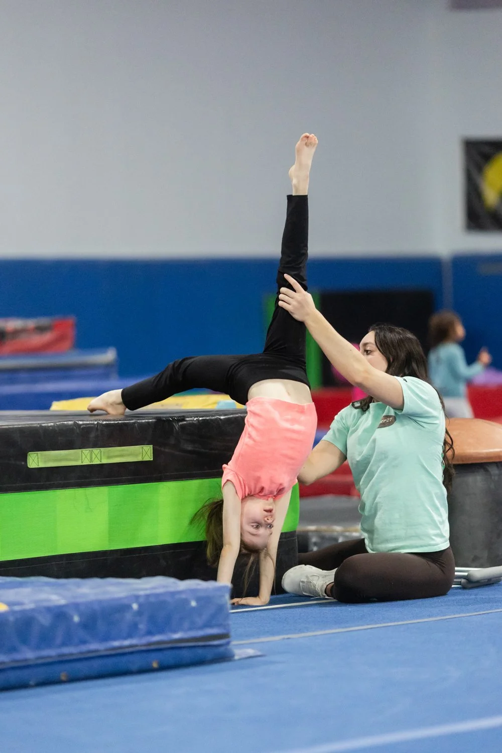 A young girl practicing gymnastics on a padded mat, with one leg extended straight up while her head and hands are on the floor, assisted by a coach during training in an indoor gym.
