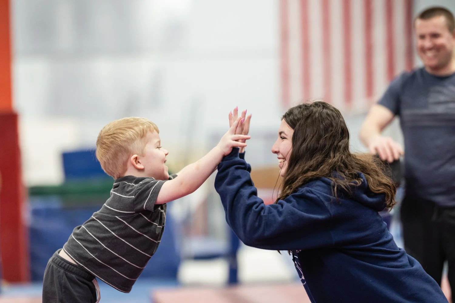 A young boy and a young woman are playing a high-five game, smiling at each other, with a man in the background smiling. They are indoors.