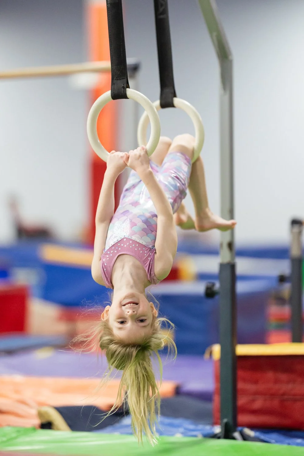 A young girl with long blonde hair hanging upside down from gymnastic rings in a gym.