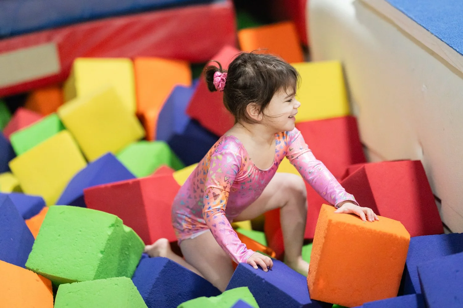 A young girl with brown hair tied in a pink scrunchie, wearing a pink long-sleeve shirt and shorts, climbing on foam blocks in a colorful indoor play area.