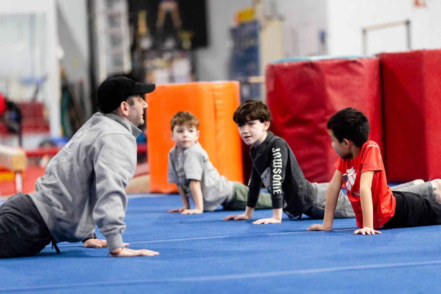 An instructor and three young boys in a gymnasium practicing push-up exercises on a blue mat, with large red and orange gym mats in the background.