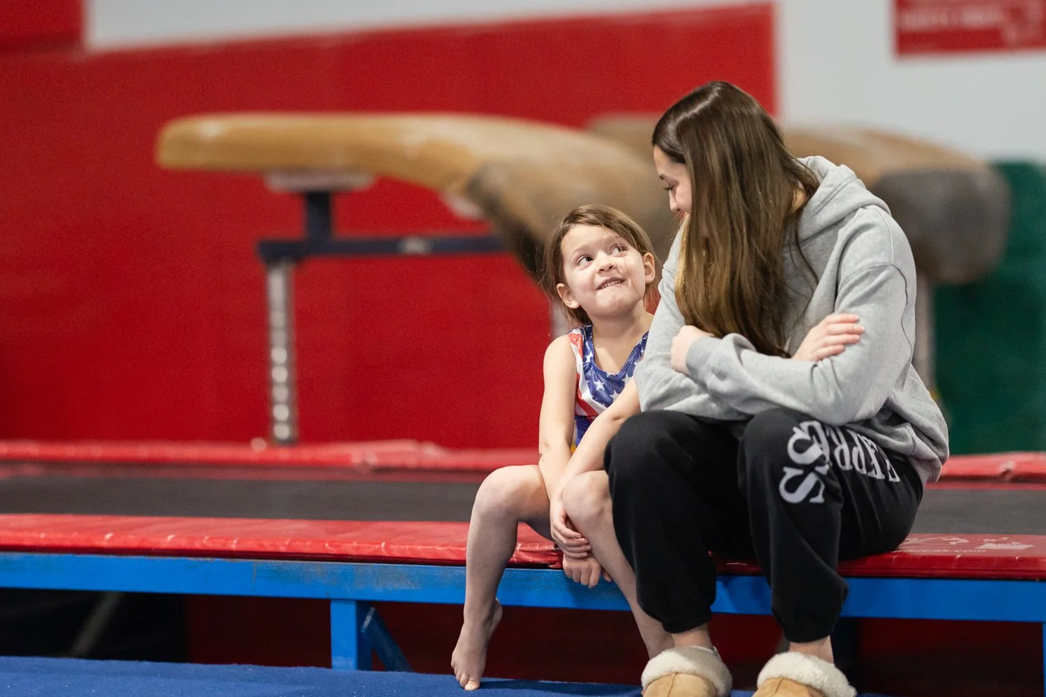 A young girl and a woman, likely her mother, sitting together on a red trampoline. The girl is smiling up at the woman, who is leaning in towards her with a smile. Both are dressed casually, and the background shows gymnastic equipment and a red wall.
