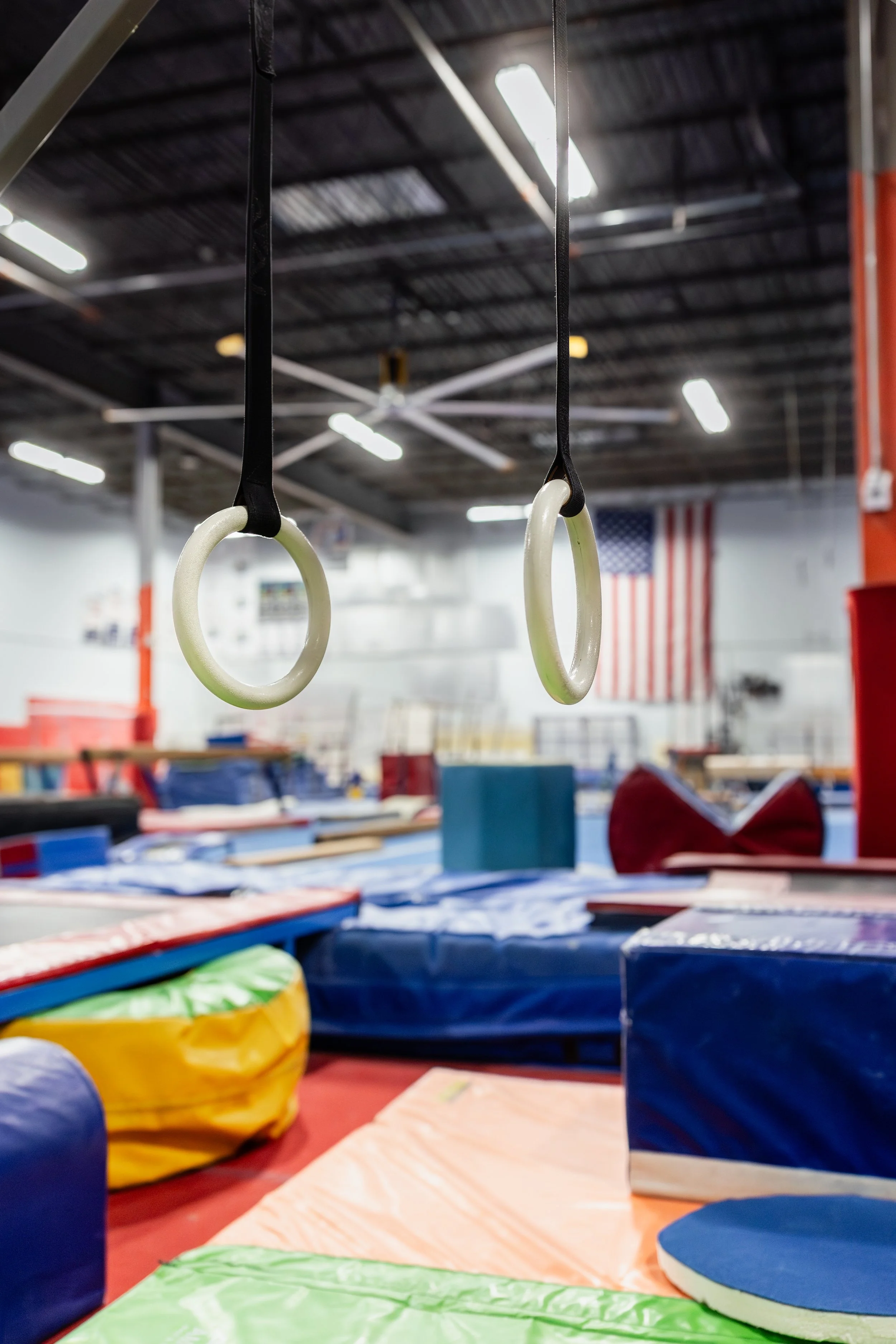 Gymnastics training area with gymnastics rings hanging from the ceiling, various mats, and other equipment in the background.