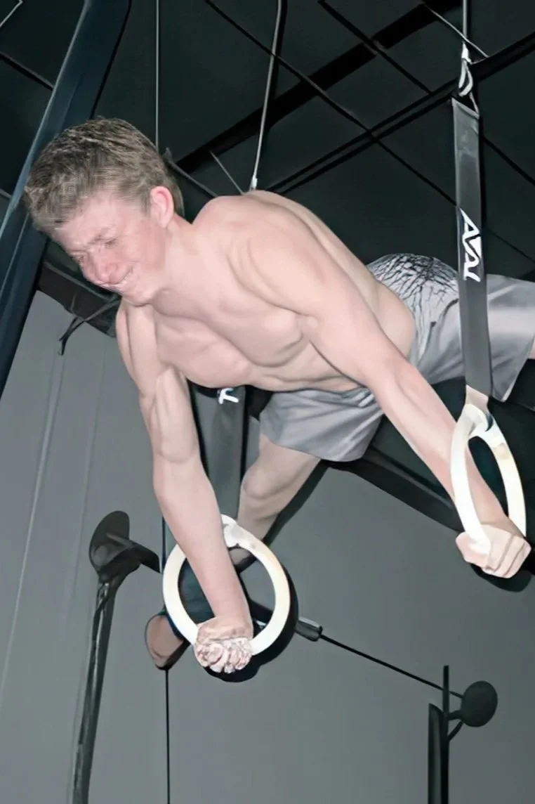 A man performing an aerial acrobatics on gymnastic rings, hanging from the ceiling of a gym or fitness studio.