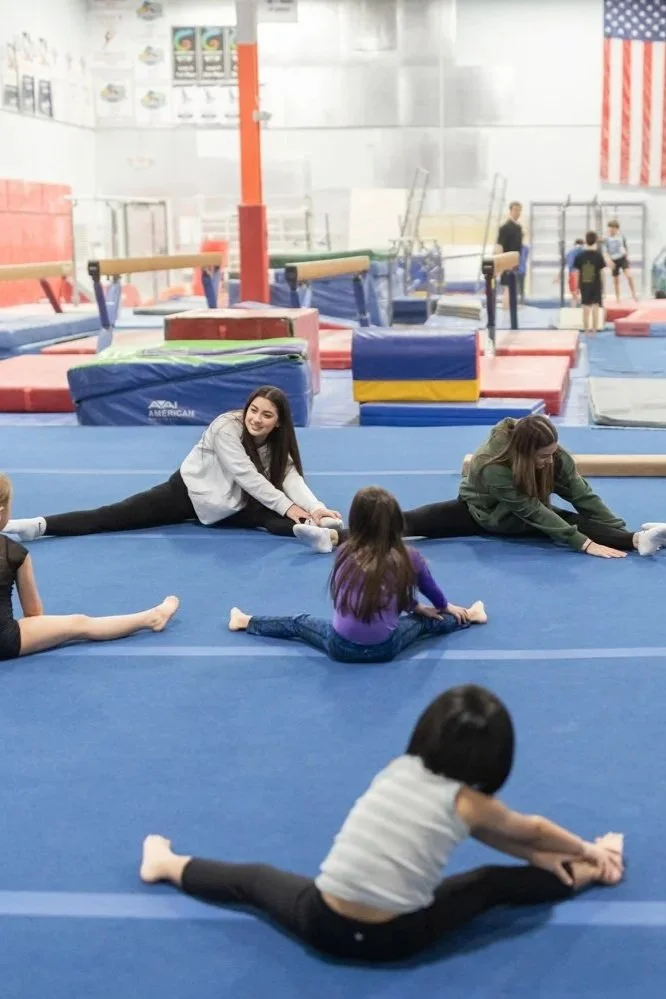 Children and women practicing gymnastics stretches on blue mats at an indoor gym. Four females are doing splits, and a young girl is sitting on her ankles in a butterfly stretch. Gymnastics equipment and an American flag are visible in the background.