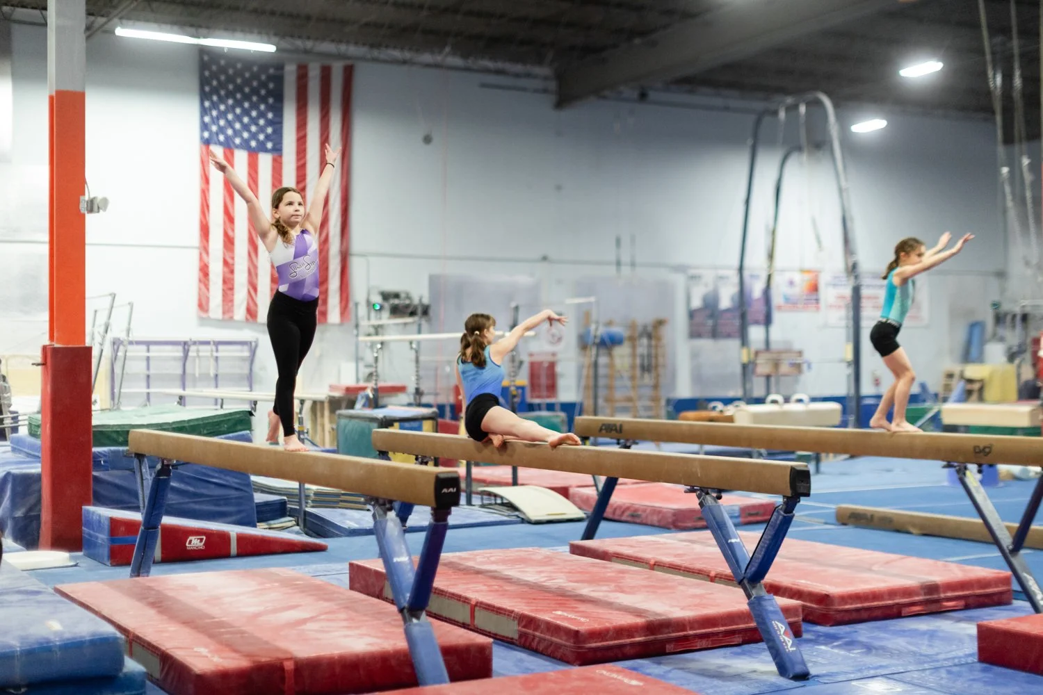 Three young girls practicing gymnastics on balance beams inside a gymnasium with an American flag in the background.