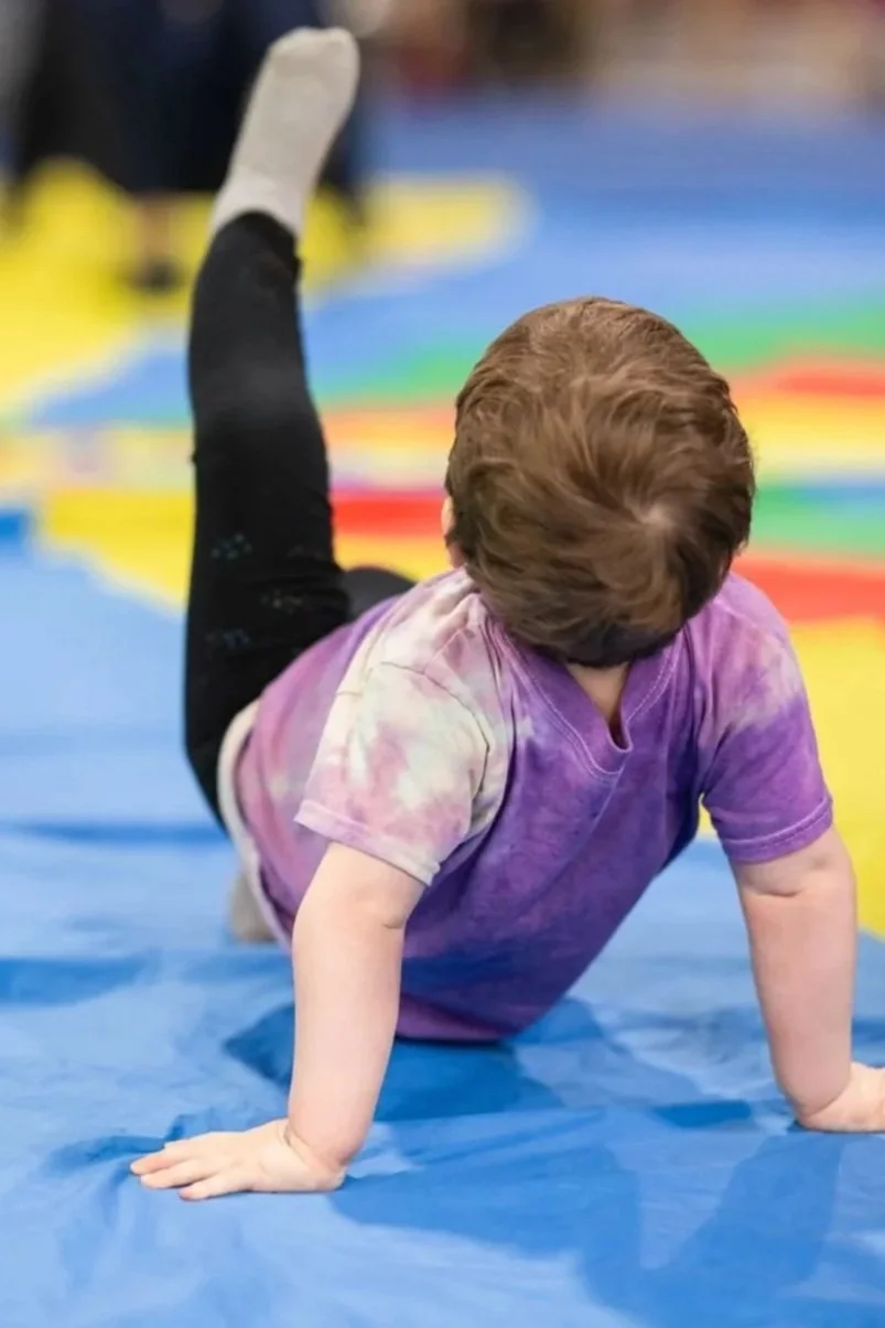 Child with brown hair in a purple tie-dye shirt and black pants performs a gymnastic move on a blue mat in an indoor gym or activity center.