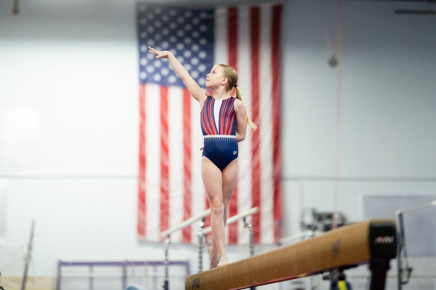 Young girl in gymnastics attire balancing on a beam in an indoor gym, with American flag in the background.