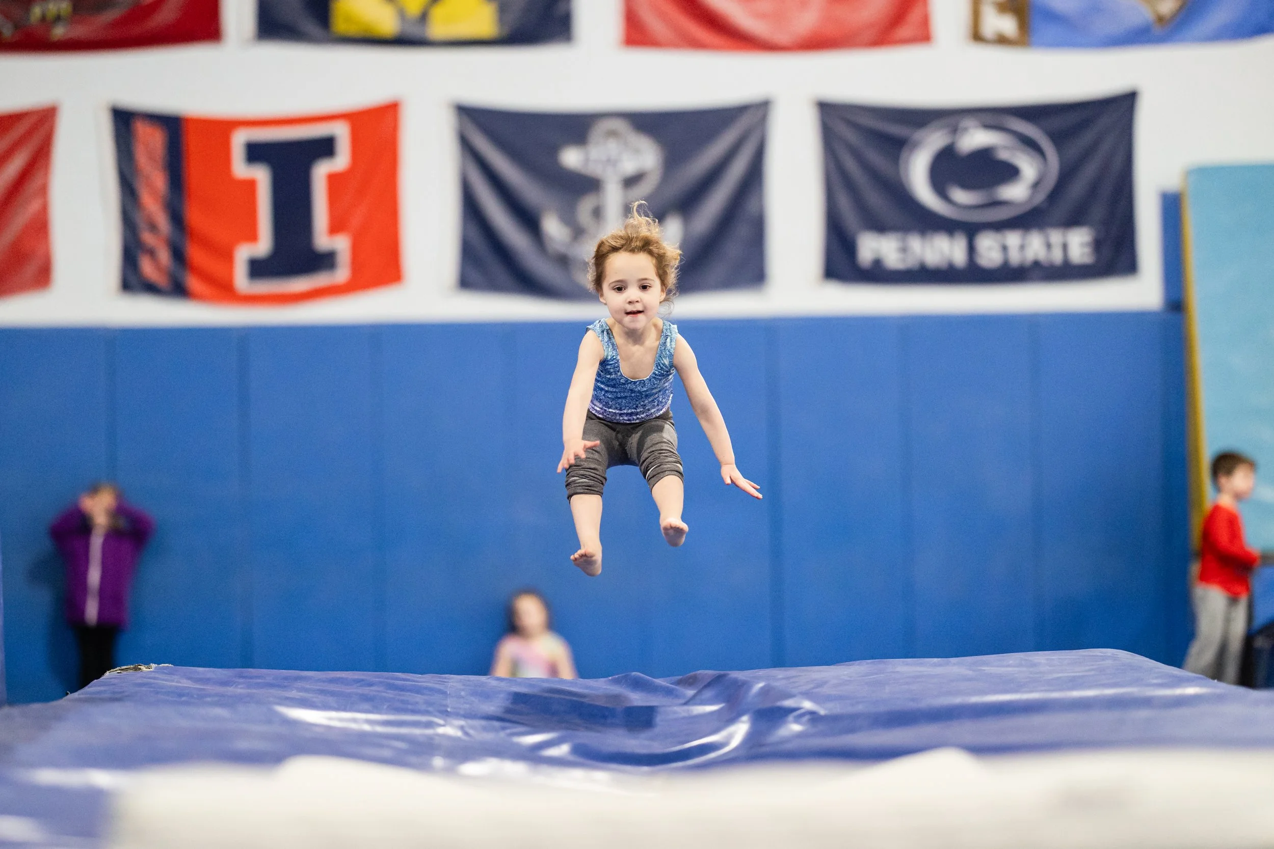 A young girl in a blue sleeveless top and gray pants jumping on a gymnastics trampoline in an indoor gymnasium, with college gymnastics banners and other children in the background.