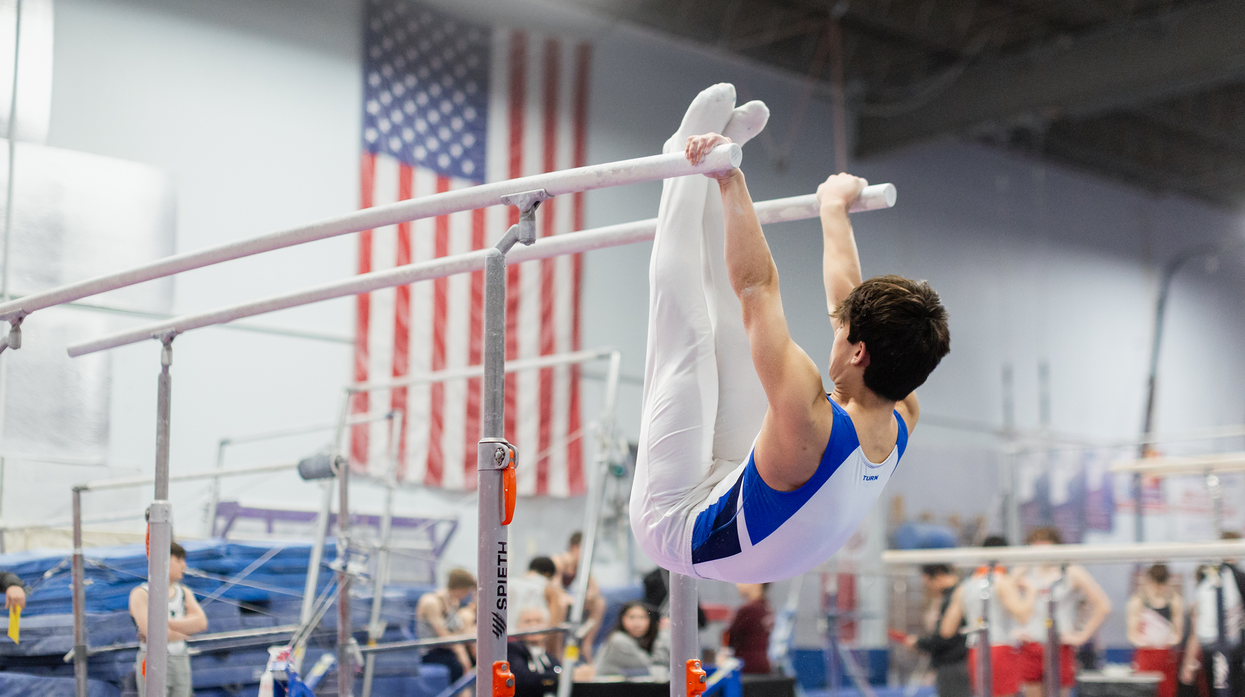 A male gymnast is hanging upside down on uneven bars, holding onto the bar with both hands inside a gymnasium with an American flag hanging on the wall.