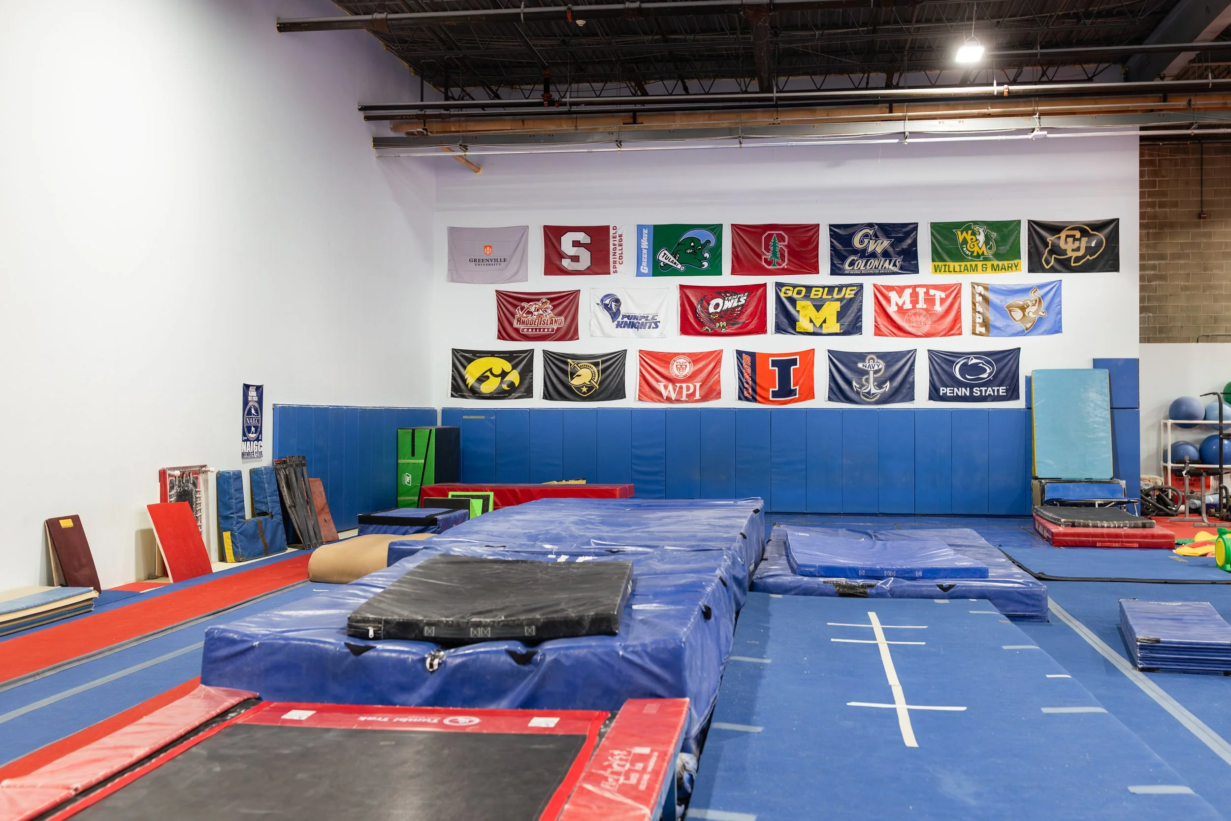 Inside a gymnastics or training facility with padded mats and walls decorated with college flags, including Greenville University, Syracuse College, Tulane, Stanford, Georgia Southern, William & Mary, and others.