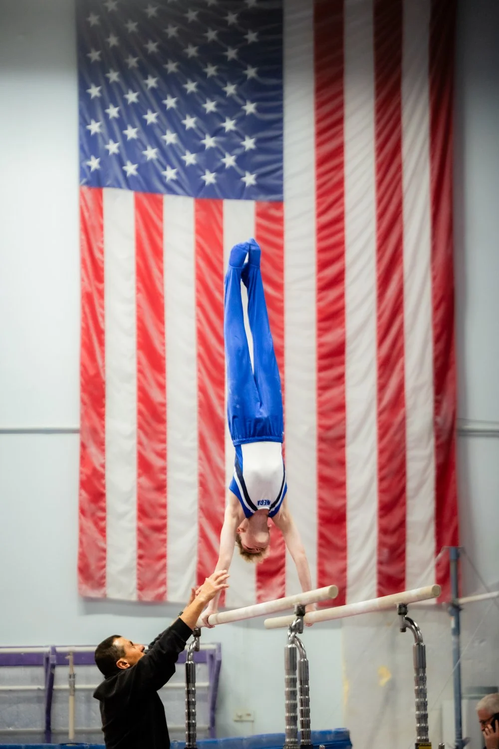 boy on parallel bars at a gymnastics meet in front of an american flag with his coach spotting him