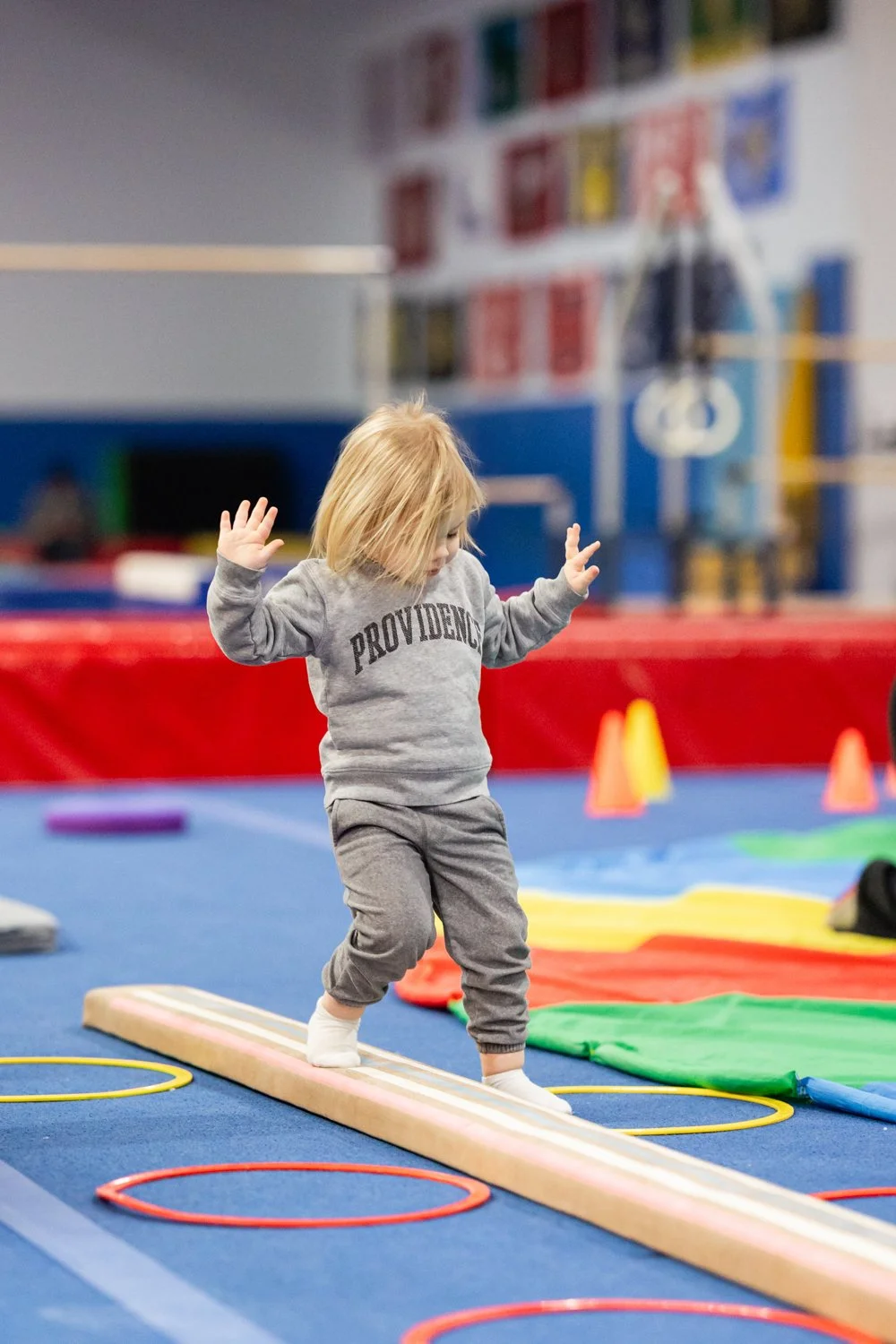 Young child with blonde hair balancing on a wooden beam in a colorful indoor play area, surrounded by various gym equipment and soft play mats.