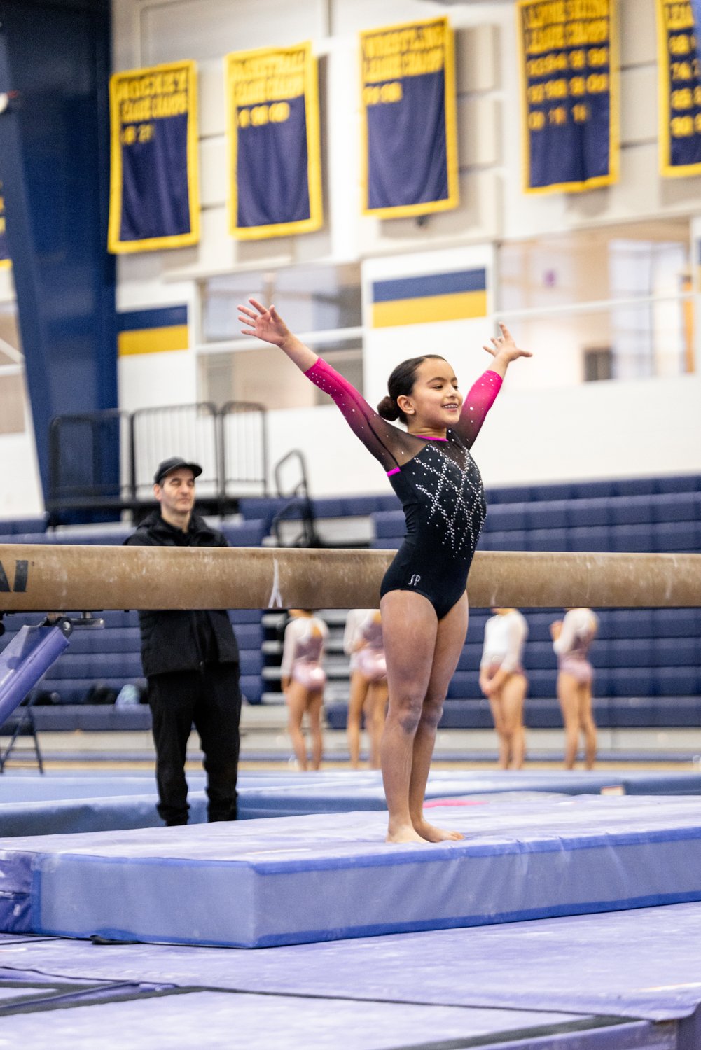 A young female gymnast stands on a balance beam with arms extended, smiling, during a gymnastics practice or competition in an indoor gymnasium. In the background, people and other gymnasts are present; yellow and blue banners hang on the wall.