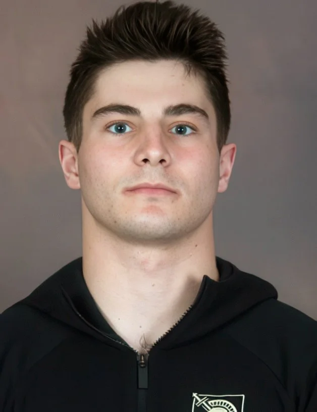 A young man with short, dark hair and blue eyes wearing a black zip-up jacket against a plain gray background.