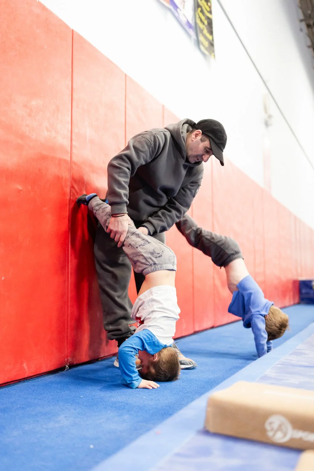 A coach assisting two young boys with handstands at a gym.