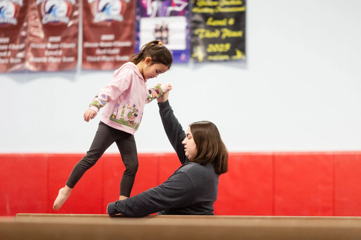 A young girl in a pink hoodie with colorful characters on it is balancing on a gym mat while a woman supports her by holding her hand in a gymnastics or cheerleading practice.