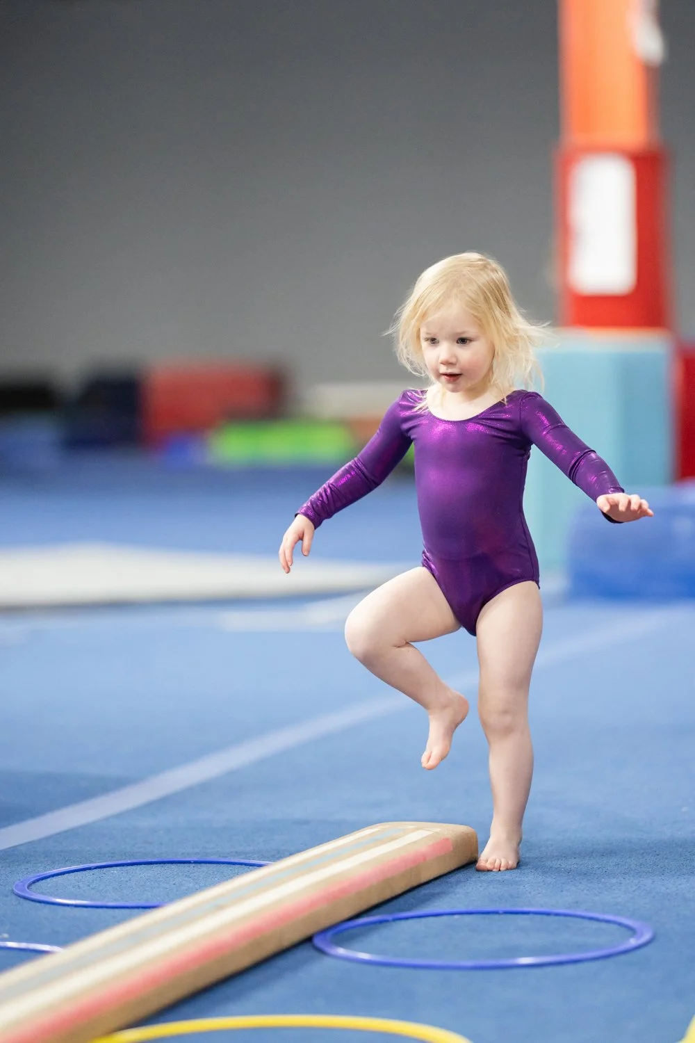 Young girl in a purple gymnastics leotard balancing on a beam in a gymnastics training facility.