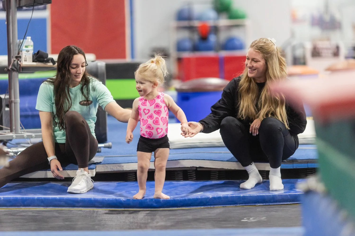 Two women and a young girl with blond hair at a trampoline gym. The girl is standing on a trampoline mat in pink and black athletic clothes, holding hands with one woman and touching the other's hand. The women are kneeling or squatting on the trampo