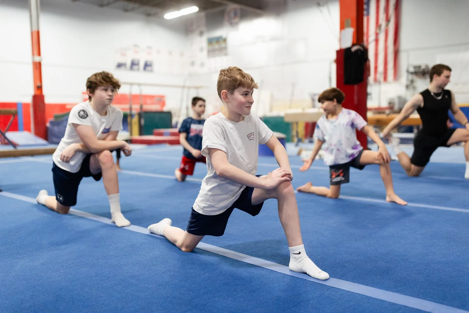 Children in a gym class practicing stretching exercises on a blue mat, with one boy in the foreground kneeling on one knee and others performing similar stretches in the background.