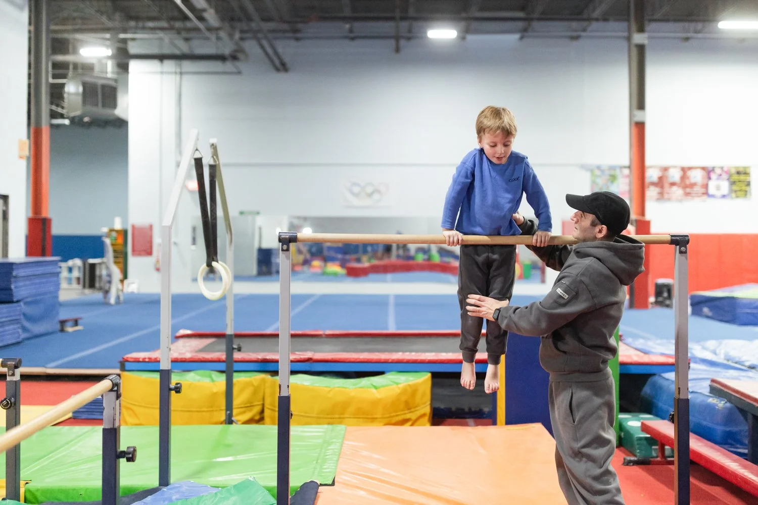 An indoor gymnastics facility with a young boy practicing with a coach. The boy is on a balance beam, and the coach is supporting him by holding him steady. The background includes mats and gymnastic equipment.