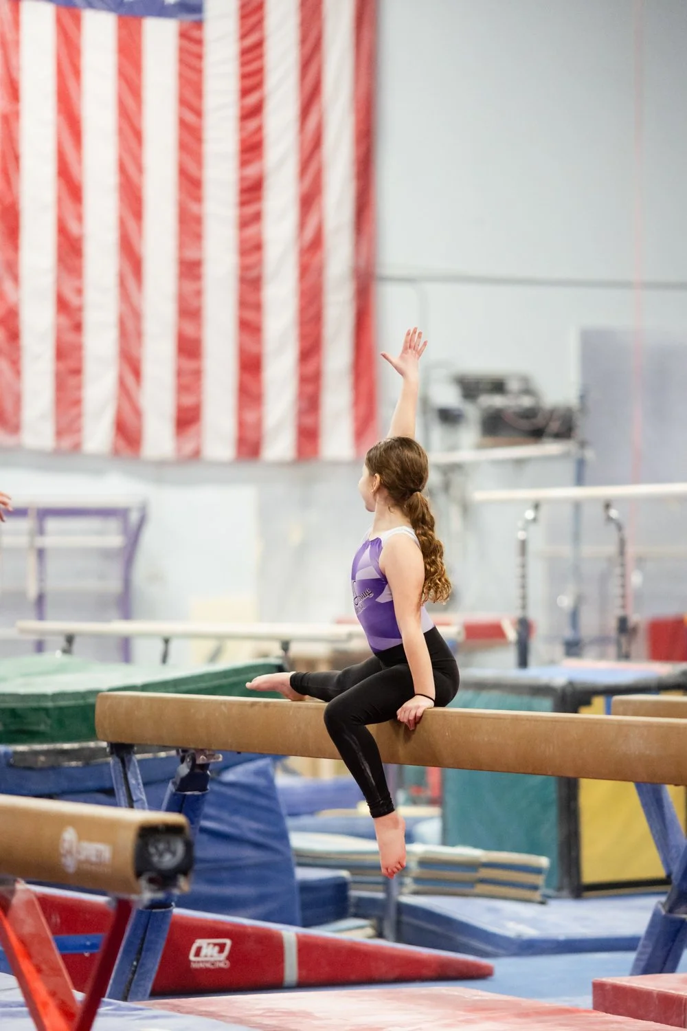 A young girl with long curly hair in a gymnastics facility, sitting on a balance beam with her right arm raised, wearing a purple and white leotard and black leggings.